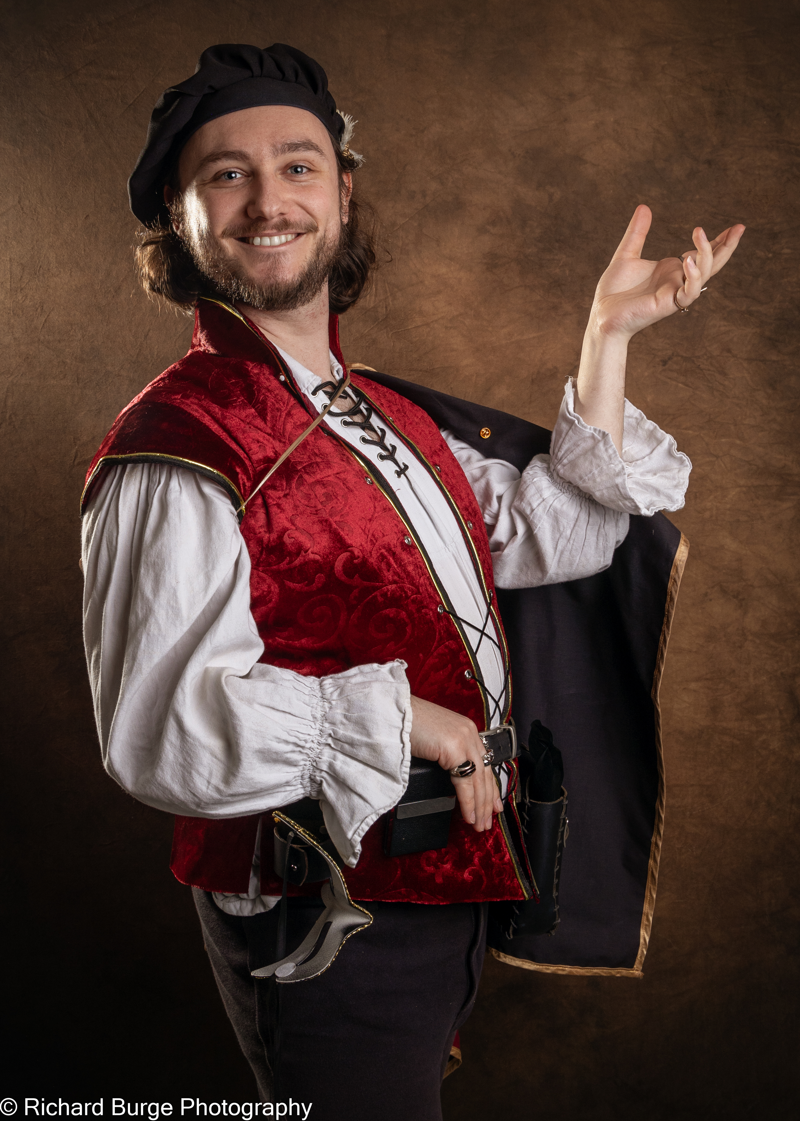 A character from the League in Empire wearing a red velvet waistcoat and shoulder cape poses with an outstretched hand and a big smile