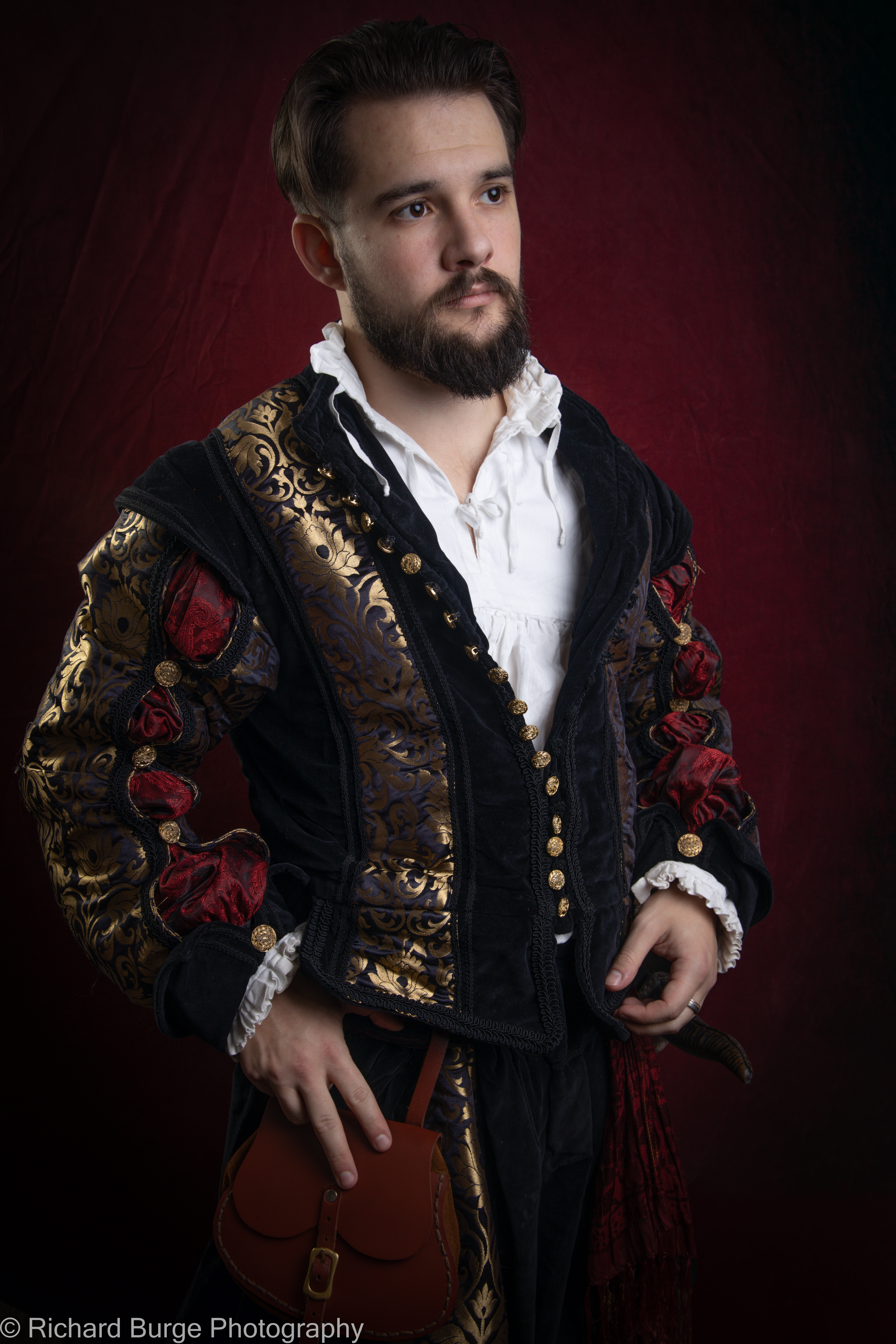 A character from the League in Empire wearing a renaissance doublet in gold and black poses against a red background gaazes thoughtfully into the distance