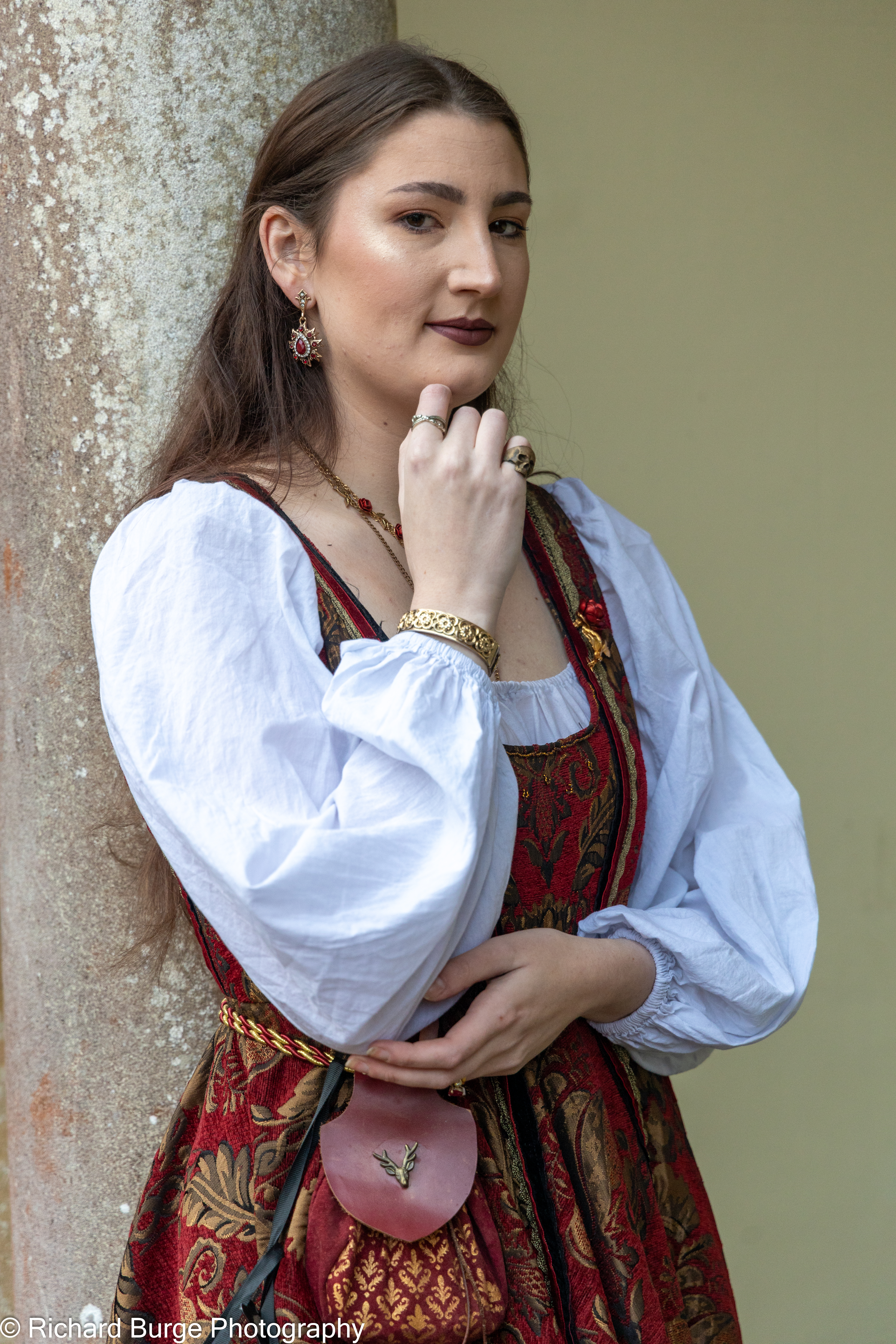 A character from the League in Empire wearing a red and gold dress over a white puffy sleeve shirt leaning against a stone wall wearing a confident thoughtful expression